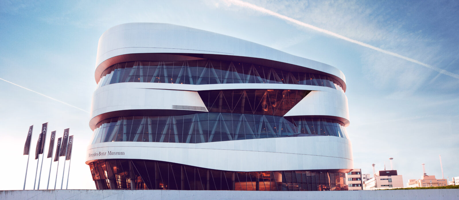 Organische Außenfassade des Mercedes-Benz Museums mit vielen Glaselementen und einem Eingangsbereich mit vorgelagertem Außenbereich, bestehend aus einem runden Vorplatz mit angelagerten Sitzgelegenheiten ähnlich einem Amphitheater.